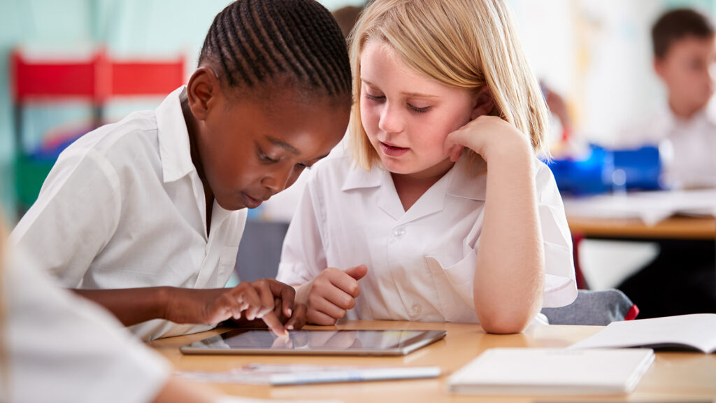 Two Elementary School Pupils Wearing Uniform Using Digital Tablet At Desk